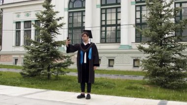 Şapkalı ve cüppeli modern mezun çocuk. Genç adam, üniversiteden başarılı bir şekilde mezun olan öğrencilerin mezuniyet töreninden birkaç dakika sonra aileleri ve arkadaşlarıyla konuşuyor.