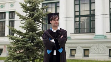 Handsome dark-haired graduate in a cap and black robe, confident and proud, nods demonstratively as a sign that he has succeeded. Boy with glasses at the university graduation ceremony