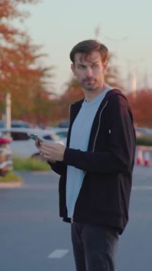 Casual-dressed young man stands in a parking lot, holding smartphone, surrounded by parked vehicles, illustrating modern carsharing accessibility and urban mobility