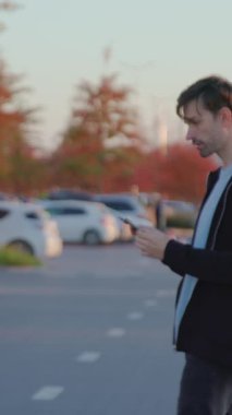 Sıradan genç bir adam park alanında yürüyor, park halindeki araçlarla çevrili akıllı telefonu kontrol ediyor, işe hazırlanıyor.