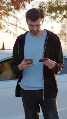 Genç adam akıllı telefonunu online ödemeler için kullanıyor. Bir kredi kartı tutuyor ve kiralık arabanın parasını ödemek için telefonda ayrıntıları giriyor. Adam internetten banka kartı ve satın alınan arabanın telefonu ile ödeme yapıyor.