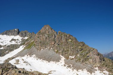 İtalya 'nın Vermiglio yakınlarındaki kayalık bir dağın çarpıcı manzarası. Manzara, parlak mavi gökyüzünün altında engebeli kayaların ve kar parçalarının kontrastını vurgular..