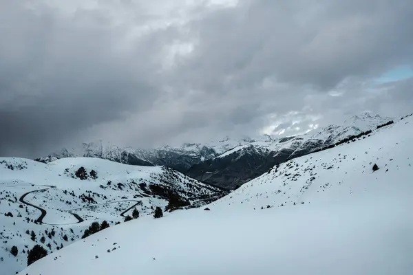 Kıvrımlı bir yolu olan karlı dağ manzarası, bulutlu gökyüzü ve bulutlarla kısmen gizlenmiş uzak dağlar. Kara kayalar ve kardan görülebilen seyrek bitki örtüsü. Sakin ve soğuk sahne.