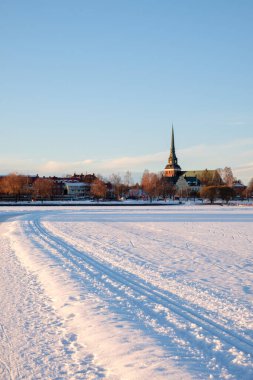 Snow blankets the ground in Mora, Dalarna during winter, creating a serene atmosphere. Trails lead towards a charming town with a distinctive church spire in the background. clipart