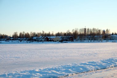 In winter, a serene snowy expanse stretches across Mora, Dalarna, revealing distant buildings and trees against a clear sky. This scene captures the beauty of a chilly day by the lakeside. clipart