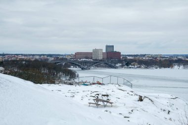 Stockholm 'de güzel bir kış manzarası donmuş bir göl ve karla kaplı bir zemin gösteriyor. Ufuk çizgisinde, mevsim boyunca sakin ve serin bir atmosferi çağrıştıran binalar ve bir köprü yer alıyor..