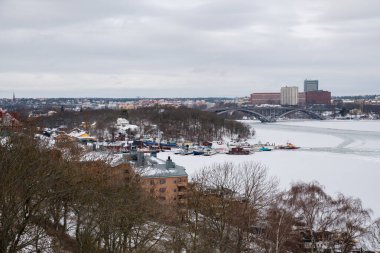 Kar Stockholm 'ün sahil şeridini kaplayarak sakin bir kış manzarası oluşturuyor. Renkli tekneler buzun arasına demirlenirken, binalar bu serin günde bir zemin oluşturuyor..