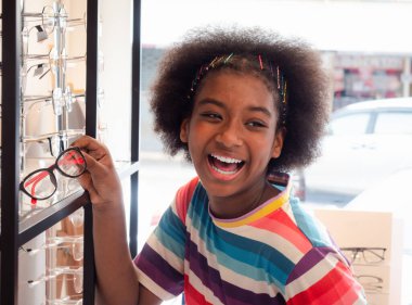 Optik mağazasındaki Afrikalı kız yeni bir gözlük çerçevesi seçmek için aynaya bakıyor. Sağlık, ilaç, insanlar, görme ve teknoloji kavramı