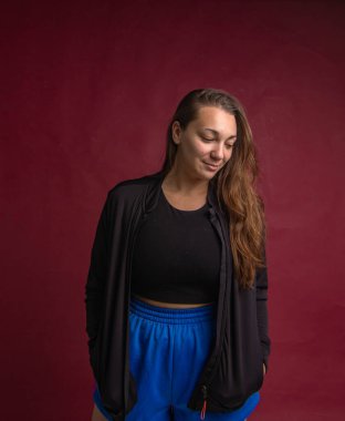 Siyah ceketli ve mavi şortlu bir kadın gülümsüyor. Kırmızı bir arka planın önünde duruyor.