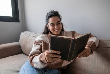 Bir kadın kanepede oturmuş kitap okuyor. Gülümsüyor ve eğleniyor.