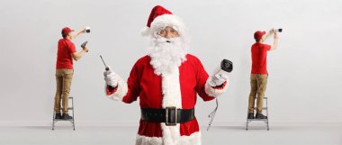 Güvenlik kameraları yerleştiren teknisyenler ve elinde bir kamera ve tornavida olan Noel Baba. 