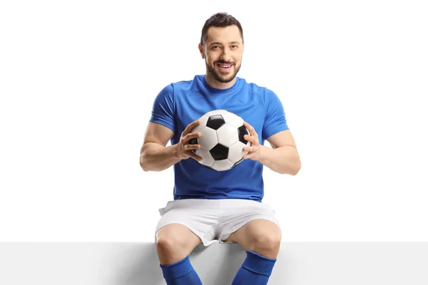 足球运动员坐在一块空白的板子上 拿着一个被白色背景隔开的足球 — 图库照片