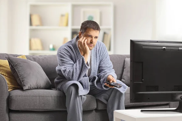 若いです男でバスローブを着てテレビを見ますと座っていますソファに家 — ストック写真