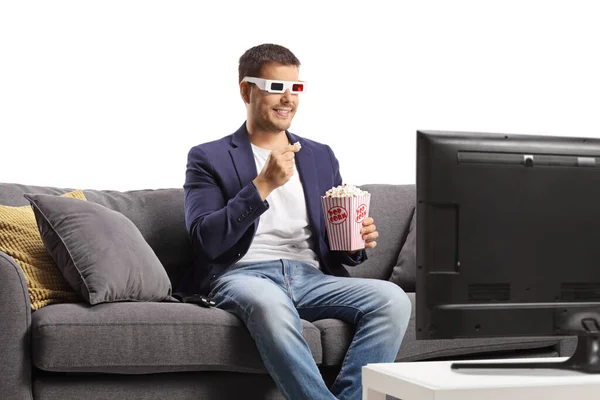 年轻面带微笑的男人 戴着3D眼镜 一边吃爆米花 一边坐在家里的沙发上观看白色背景的电视 — 图库照片