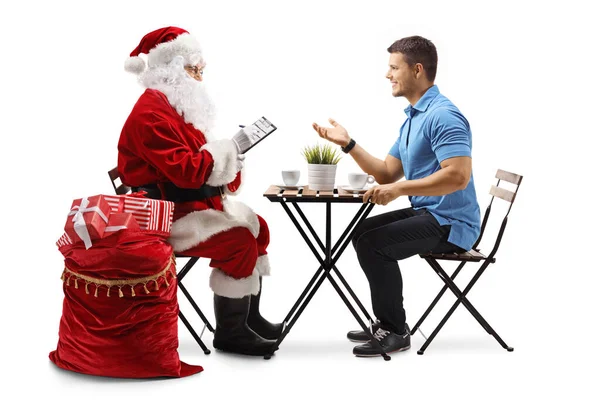 圣诞老人在写一份名单 而年轻人则坐在一间被白色背景隔开的咖啡馆里聊天 — 图库照片