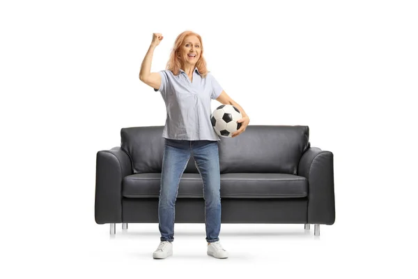 一个成熟的女人 拿着足球 在被白色背景隔开的黑色沙发前欢呼 — 图库照片