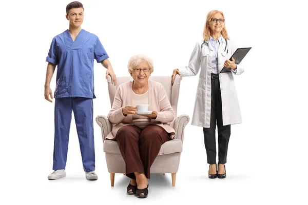 女医生和男护士站在一位坐在扶手椅上的老年妇女旁边 手里拿着一杯与白色背景隔离的茶 — 图库照片