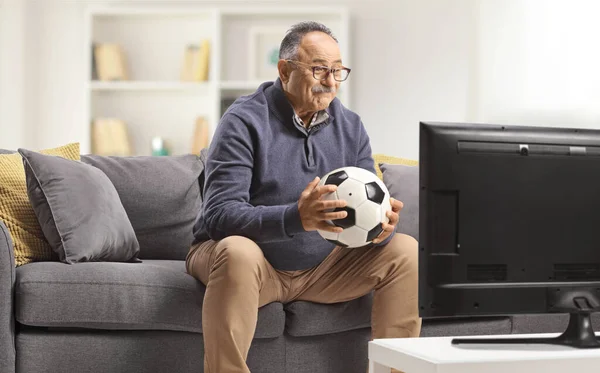 Starší Muž Sedí Pohovce Drží Míč Dívá Fotbal Televizi Doma — Stock fotografie