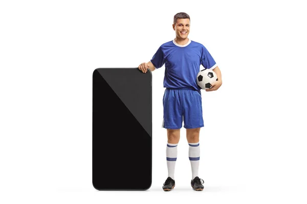 白い背景に隔離された大きなスマートフォンの隣に立っているサッカージャージの男 — ストック写真