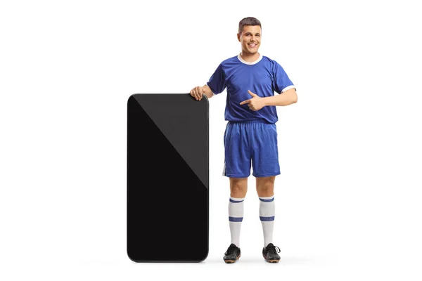 穿着足球球衣的男人站在一个大智能手机旁边 指着被白色背景隔离的屏幕 — 图库照片