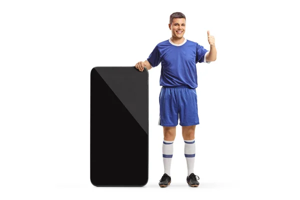 穿着足球球衣的男人站在一个大智能手机旁边 用白色的背景把大拇指伸出来 — 图库照片