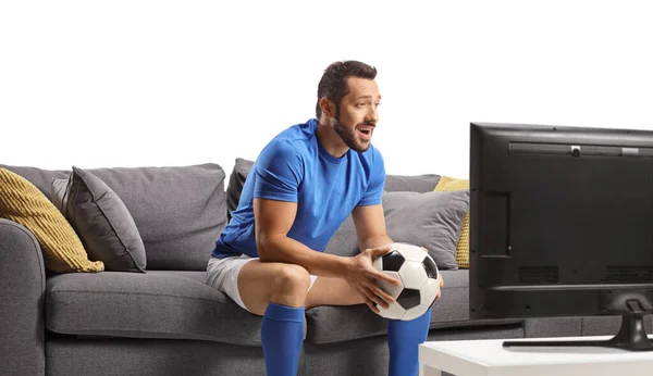 Młody Człowiek Sportowej Koszulce Siedzi Kanapie Trzymając Piłkę Oglądając Piłkę — Zdjęcie stockowe