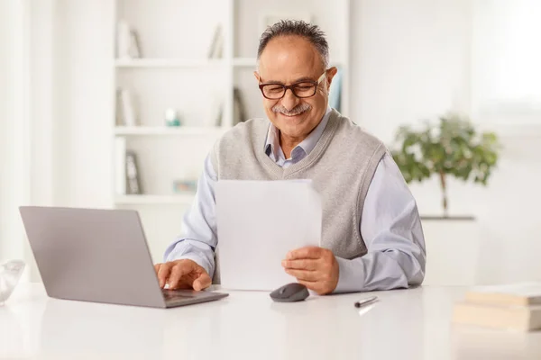 Uśmiechnięty Dojrzały Mężczyzna Trzymający Papierowy Dokument Siedzący Przed Laptopem Domu — Zdjęcie stockowe
