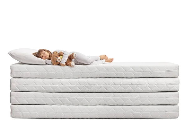 穿着睡衣的小女孩抱着一只泰迪熊睡在一堆被白色背景隔开的床垫上 — 图库照片