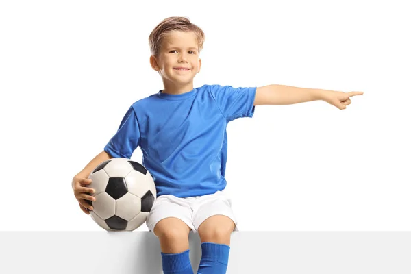 身穿蓝色球衣的男孩坐在一块空板子上踢足球 指着被白色背景隔开的一边 — 图库照片