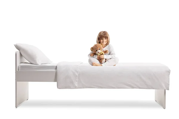 小さな女の子ですパジャマ抱擁テディベアと座っているベッドの上に隔離された白い背景 — ストック写真