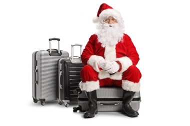 Noel Baba bir bavula oturmuş beyaz arka planda izole edilmiş kameraya bakıyor.
