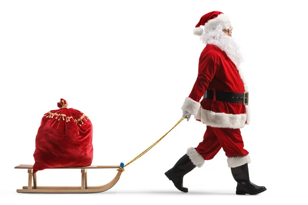 白い背景に隔離されたプレゼントでいっぱいの赤い袋で木製のそりを引くサンタクラスの完全な長さのプロフィールショット — ストック写真