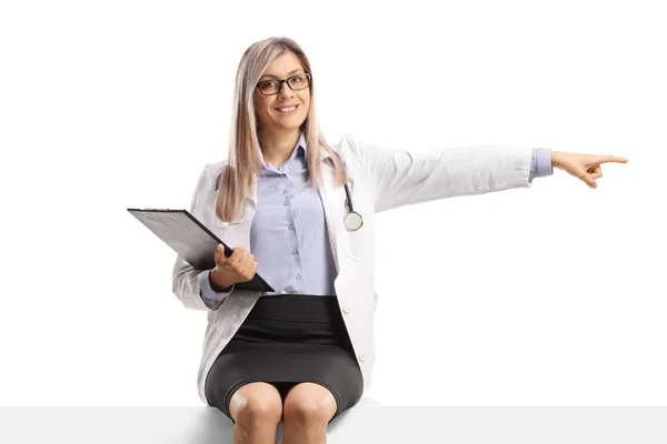 年轻的女医生坐在一块空白的板子上 指着被白色背景隔开的侧面 — 图库照片