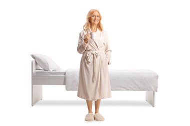 Bornozlu bir kadın elinde fincanla beyaz arka planda izole bir şekilde yatağın önünde duruyor.