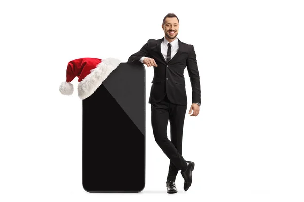 一个商人靠在一个大手机上 戴着一顶白色背景的圣诞帽的全长肖像 — 图库照片