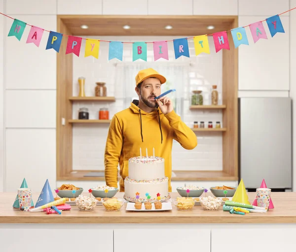 カジュアルな若い男がカウンターの後ろにパーティーホーンを吹いてケーキと好意と幸せな誕生日フラグでキッチン — ストック写真