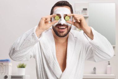 Bornozlu ve maskeli bir adam banyoda gözlerinde salatalık tutuyor.