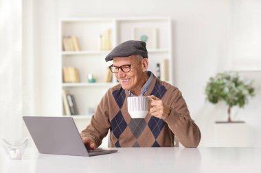 Elderly man with a cup of tea sitting at home and using a laptop computer