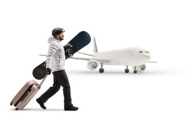 Young man holding a snowboard and pulling a suitcase in front of an airplane isolated on white background