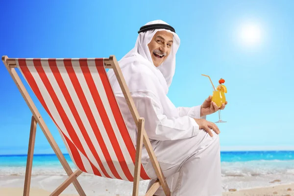 Arab man in a robe sitting on a beach with a cocktail and smiling