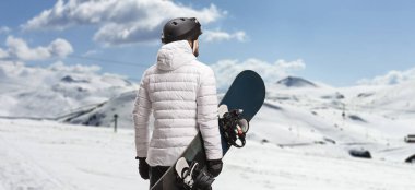 Rear view shot of a man holding a snowboard and looking at a mountain