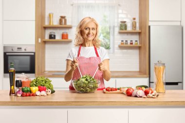 Woman behind a kitchen counter preparing a salad and smiling at camera