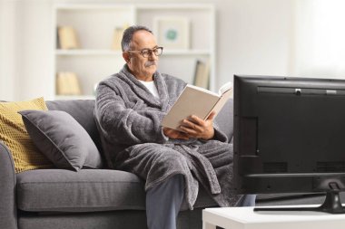 Mature man in a bathrobe sitting on a sofa in front of tv and reading a book at home