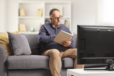 Mature man sitting on a couch and reading a book in front of tv at home