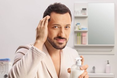Young man in a bathrobe putting on a hair conditioner inside a bathroom