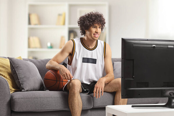 Guy in a sports jersey with a baskteball sitting on a sofa at home and watching tv 
