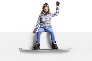 Gözlüklü, miğferli ve kar kaykaylı genç bir kadın boş bir panele oturmuş ve beyaz arka planda izole bir şekilde el sallıyor.