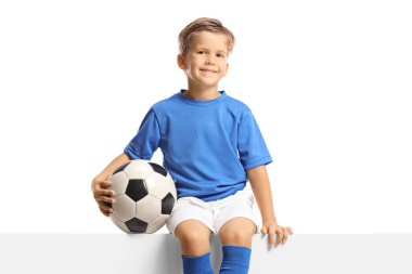 Boy in a blue soccer jersey sitting on a blank panel and holding a ball isolated on white background