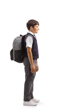 Full length profile shot of a boy in a school uniform carrying a backpack and standing isolated on white background