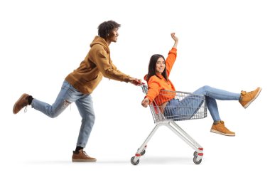 Full length profile shot of a young african american man running and pushing a girl inside a shopping cart isolated on white background